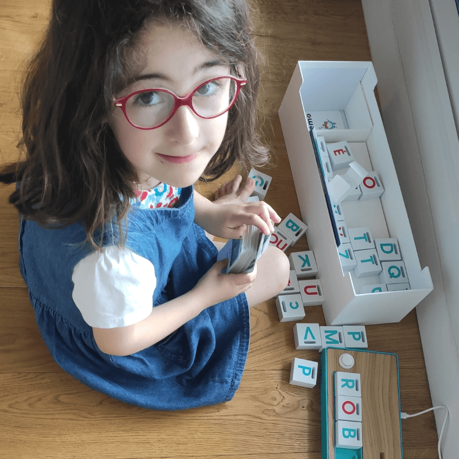Young girl with glasses sitting on the floor, engaging with colorful letter blocks scattered around her. She is holding a stack of cards and smiling, with a box of letters nearby, promoting educational play and literacy development.